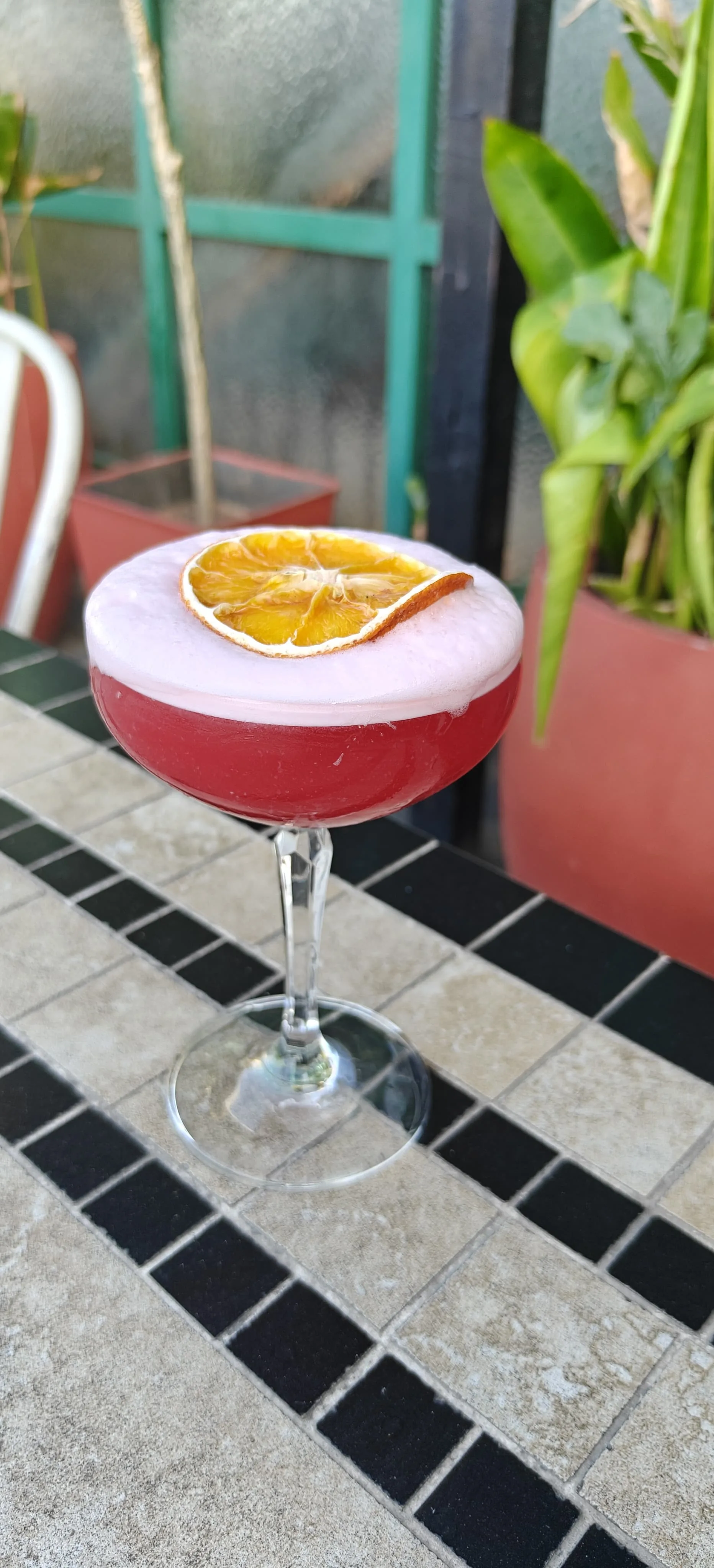 Mama's Red Lipstick — a dangerously red mezcal cocktail with a rose-pink foam cap and a dried citrus wheel, in a coupe on the rooftop